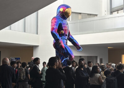 A crowd of students and visitors gathered around the tall iridescent spaceman sculpture in a modern museum atrium.