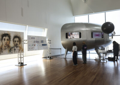 Visitors viewing fine artwork and a silver futuristic space habitat capsule inside a sunlit gallery.