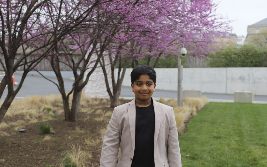 Aneeshwar Kunchala, INTERWOVEN Young Science Communicator and youth conservationist, standing outdoors in a beige suit jacket in front of blooming pink trees.