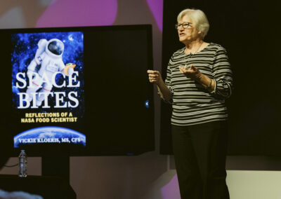 NASA food scientist Vickie Kloeris speaking next to a digital display of her book Space Bites.