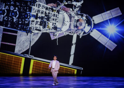 Young science communicator Aneeshwar Kunchala performing live on stage in front of a massive satellite projection.