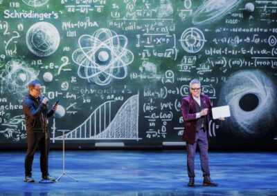 Presenters conducting a stage demonstration backed by a large chalkboard graphic of physics equations and quantum diagrams.
