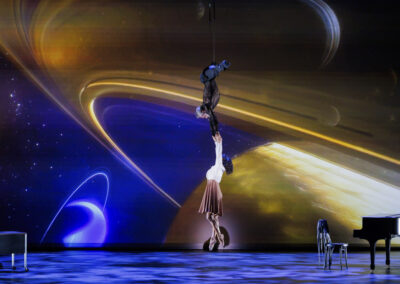 Aerial dancers suspended in mid-air during a stage performance featuring a high-resolution projection of Saturn.