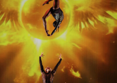 Aerial silks dancer performing high above the stage against a glowing golden sun projection.