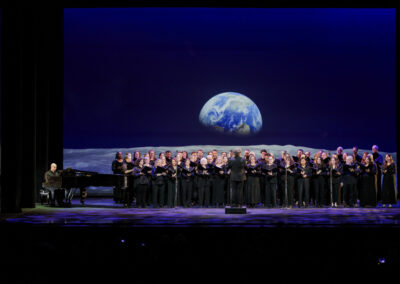 A choir and pianist performing on stage set against a dramatic projection of Earth seen from the lunar surface.