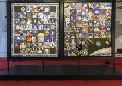 Two large museum exhibition panels displaying collaborative space-themed quilts composed of individual quilt blocks created by global community participants.