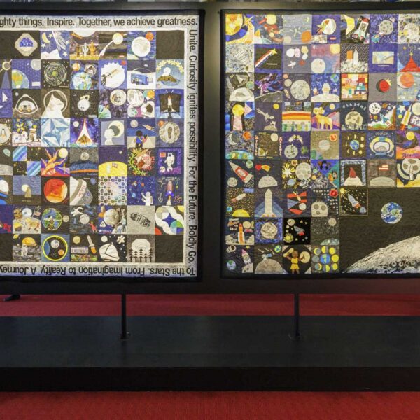 Two large museum exhibition panels displaying collaborative space-themed quilts composed of individual quilt blocks created by global community participants.