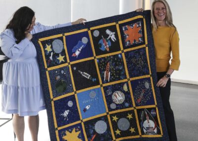Astronaut Dr. Karen Nyberg and a festival organizer holding up a custom space-themed quilt featuring rocket and planet designs inspired by NASA's Artemis campaign.
