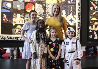 Astronaut Dr. Karen Nyberg posing with young children, including one in a NASA spacesuit costume, in front of the massive community-sourced Lunar Quilts.