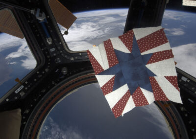 A red, white, and blue star quilt block crafted by astronaut Karen Nyberg floating in microgravity inside the International Space Station Cupola, with Earth visible below.
