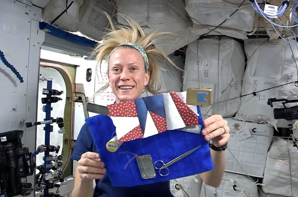 Astronaut Dr. Karen Nyberg floating in microgravity aboard the International Space Station with her hair floating upwards, holding a handcrafted red, white, and blue star quilt block.