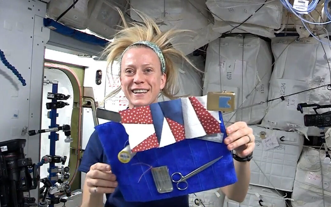 Astronaut Dr. Karen Nyberg floating in microgravity aboard the International Space Station with her hair floating upwards, holding a handcrafted red, white, and blue star quilt block.